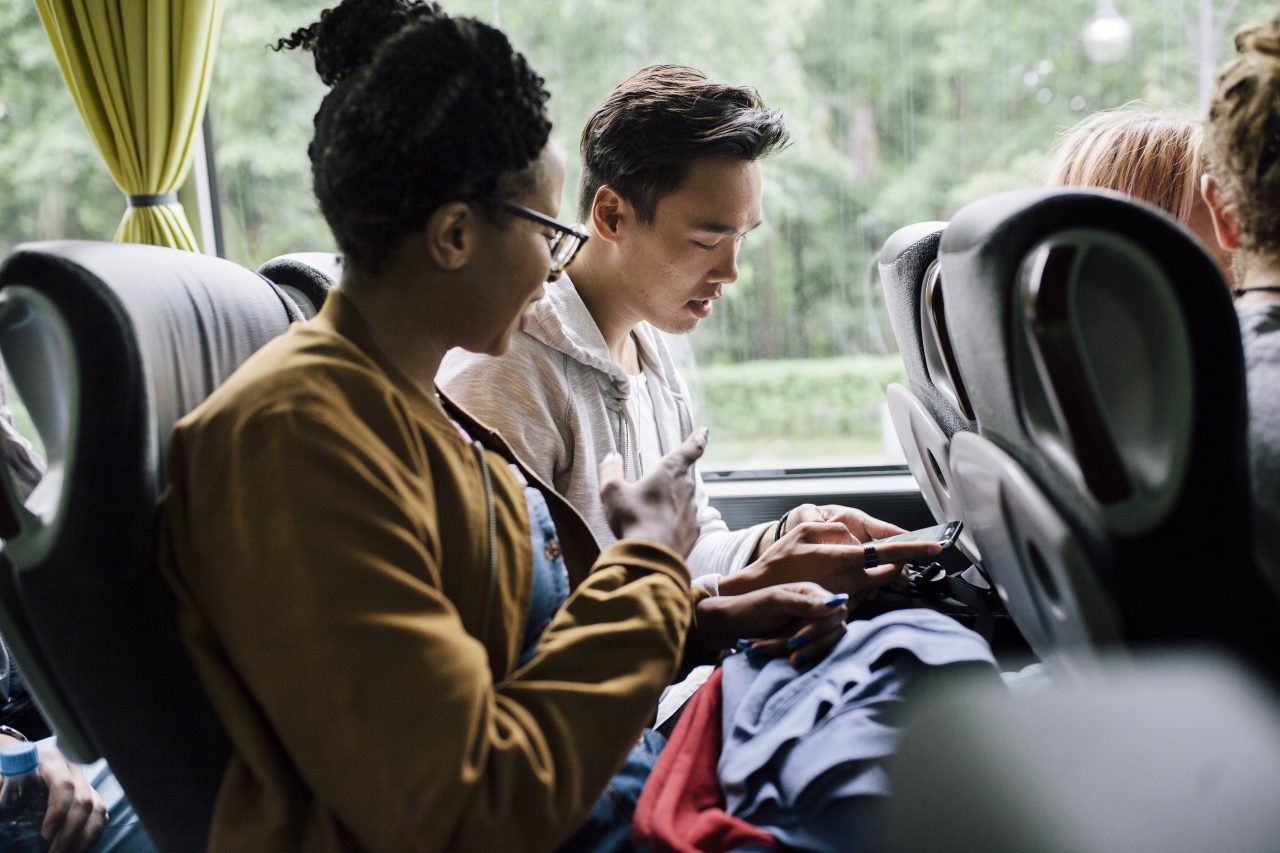 Two young friends are chatting and socialising using their smartphones as they travel together on a bus trip.